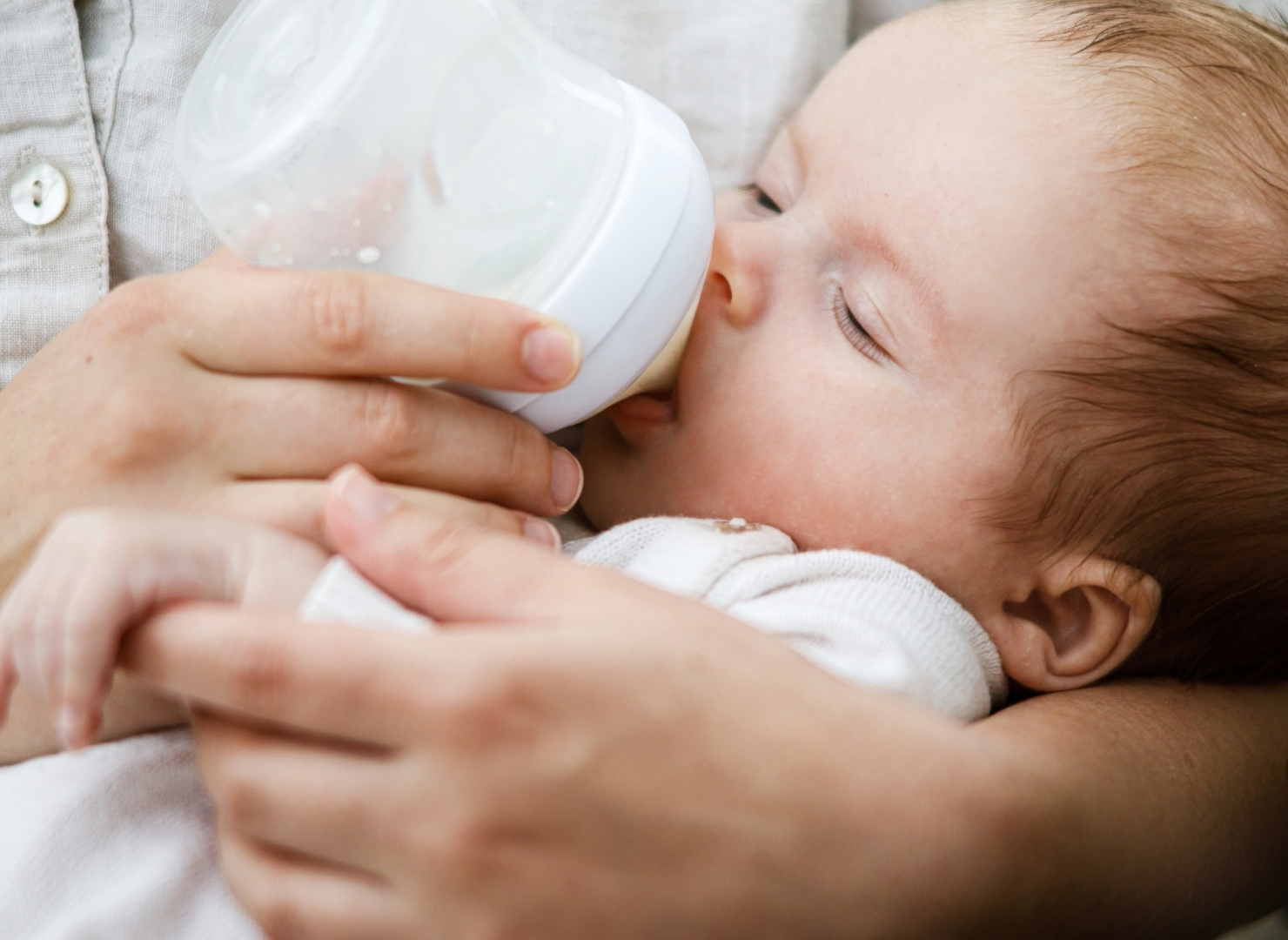 A mother holds a baby close and gently feeds from a white bottle, with the baby resting with eyes closed on soft bedding.