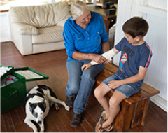 A woman wrapping a bandage around a young boys arm while sitting on a wooden bench, accompanied by a dog,