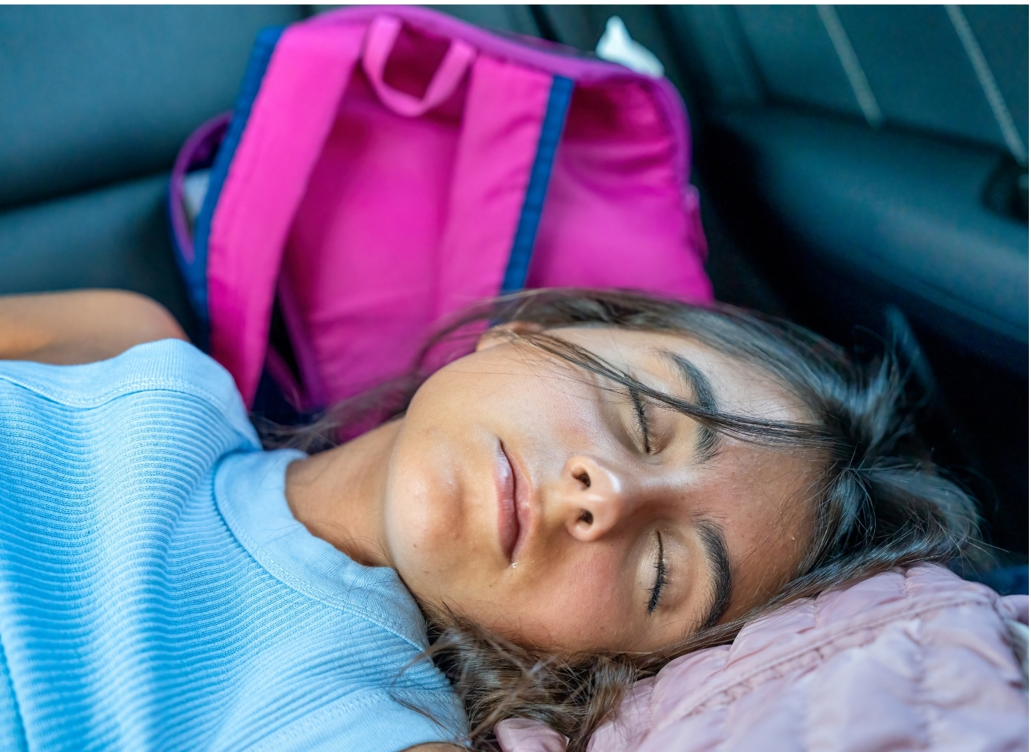 A person girl is sleeping in the backseat of a car, wearing a light blue shirt, with a bright pink bag in the background.