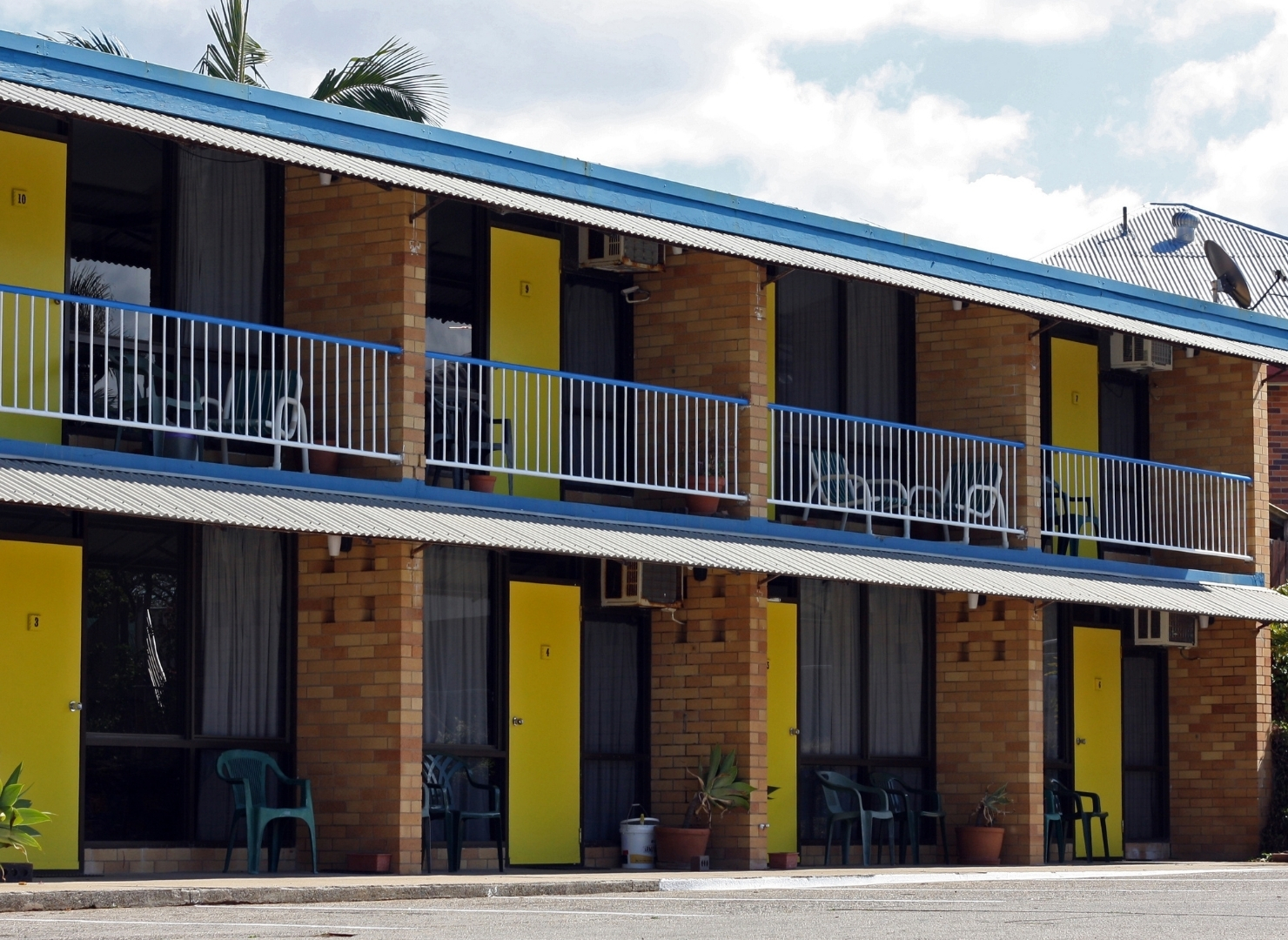 Two-story brick motel building with blue metal railings and a corrugated roof, featuring yellow doors on each level, black window shutters, and several chairs and potted plants along the front.